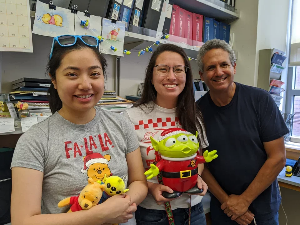 Two lab members hold stuffed toys in lab - a pooh bear and alien from the movie Toy Story - Dr. Teitell leans in next to them, smiling