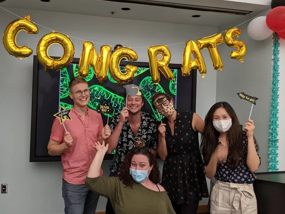 five people gather under balloons that spell "congrats" in celebration of a grad student's thesis defense