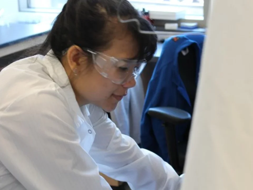 A student in a lab coat and safety goggles leans over, reaching into a hood