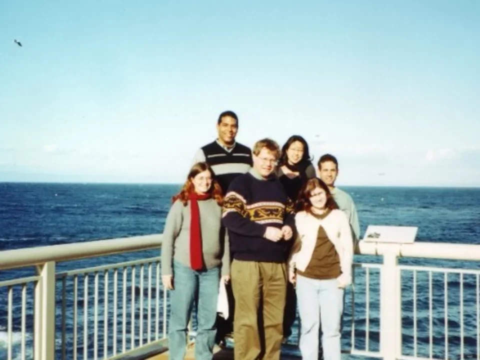 A photo from 2000 of the early Teitell Lab on a trip. Six lab members cluster next to a railing on a pier by the ocean. 